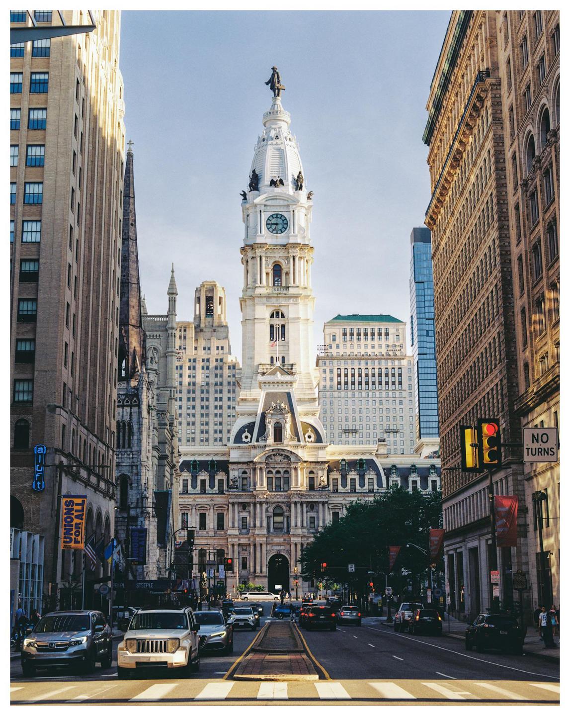 A street in Philadelphia leads to a large, ornate building with a clock tower and a statue on top.
