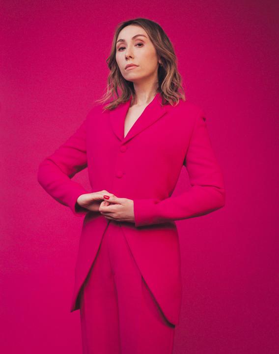A woman in a pink suit stands against a pink background with her hands clasped.