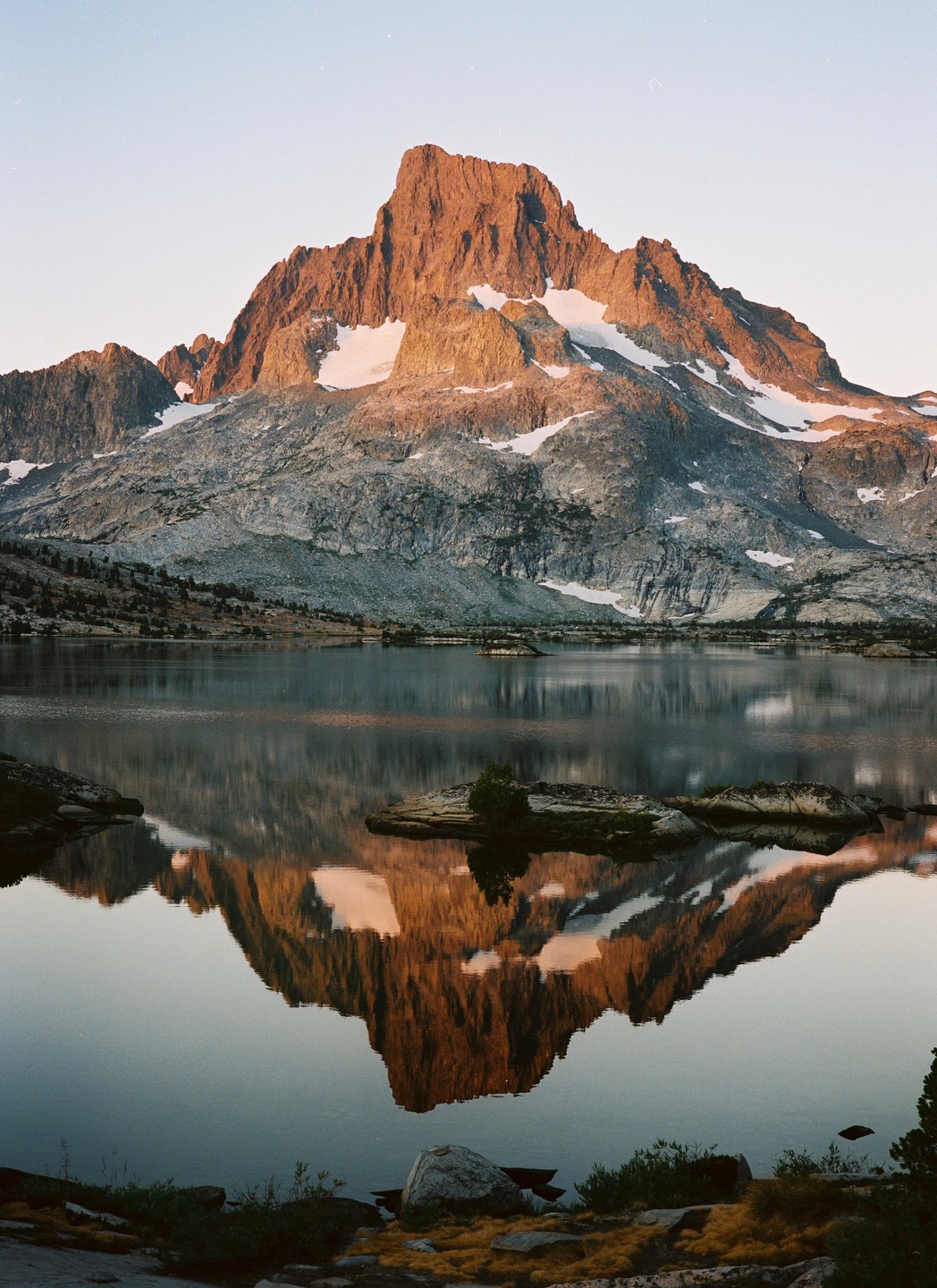 A large mountain with snow and rock is reflected in a still lake at sunset.