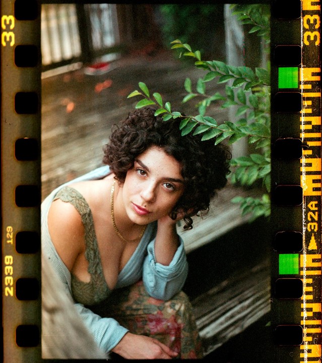 A woman with curly hair and a gold necklace is sitting on a wooden surface with green foliage in the background.