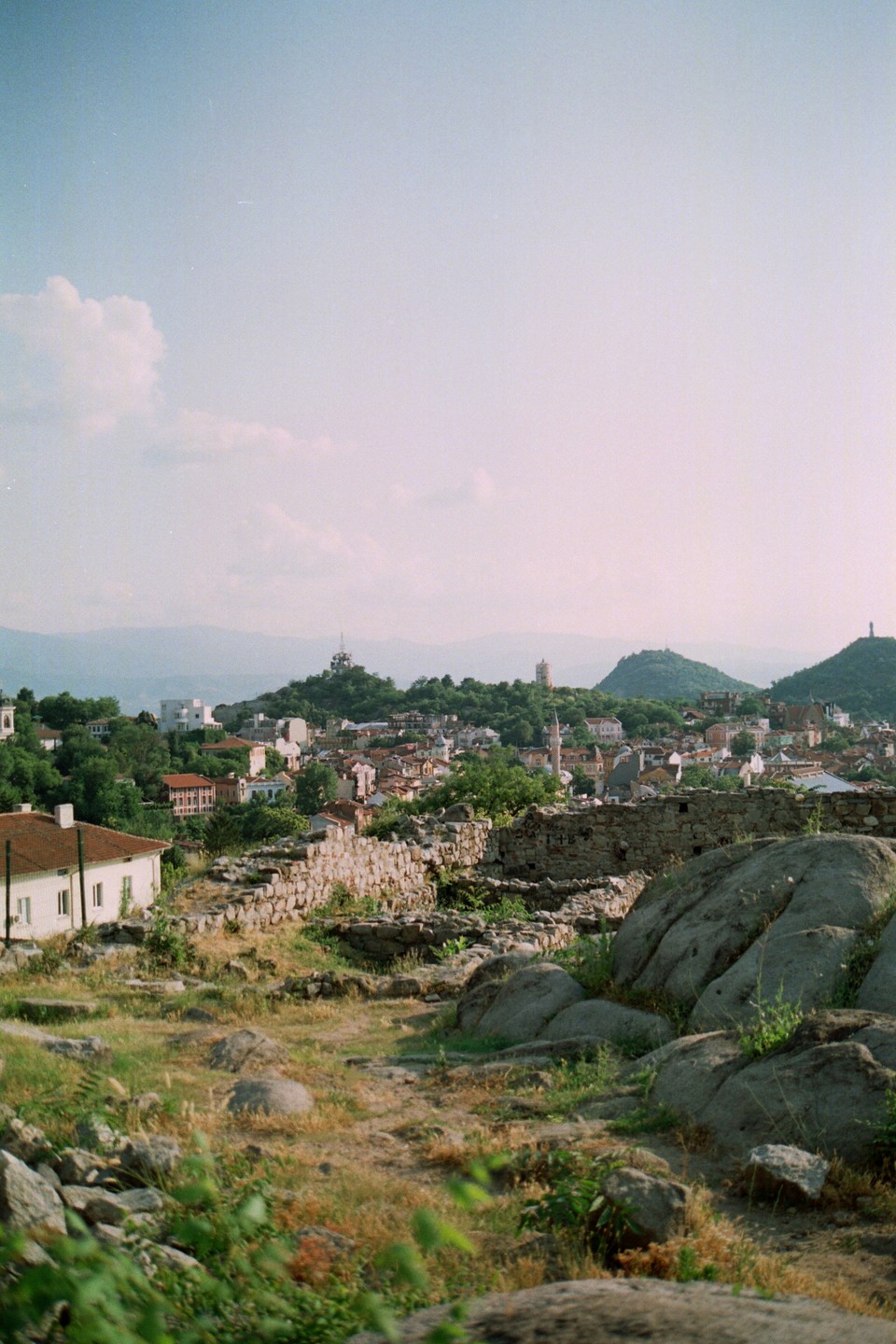 A hillside view shows a town with red-roofed buildings and green trees, with mountains in the distance under a hazy sky.