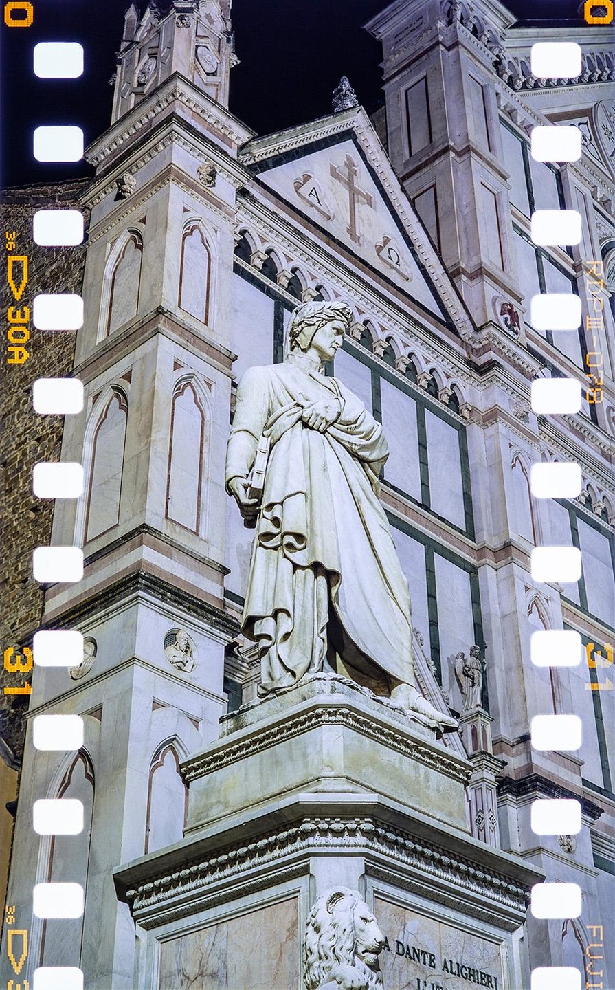 A statue of Dante Alighieri stands in front of a large building at night.