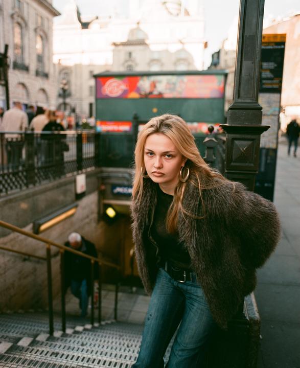 A woman in a fur coat and jeans stands on a street next to an underground entrance.