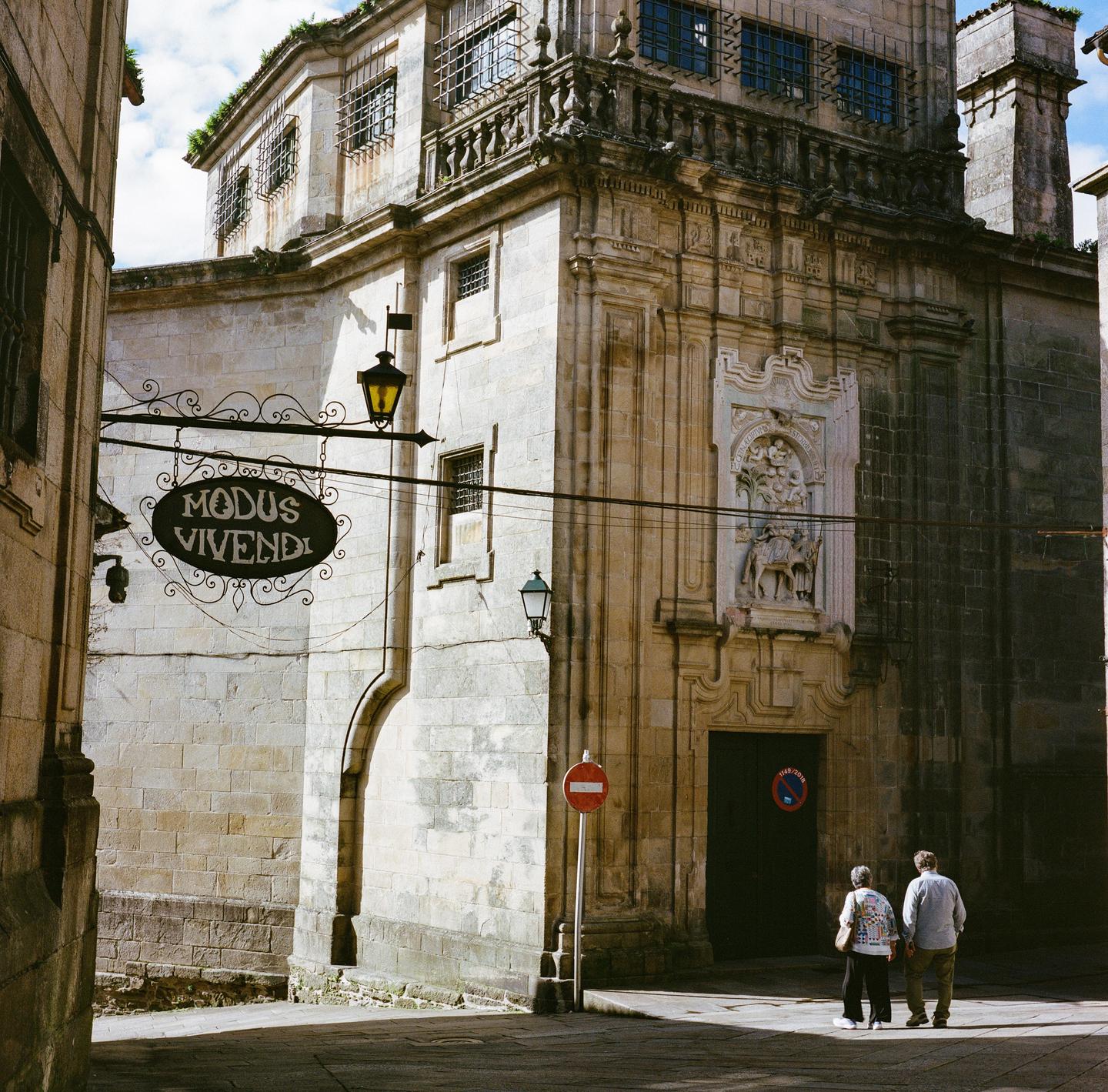 A couple stands on a street corner looking at a large stone building with a sign that reads "Modus Vivendi".