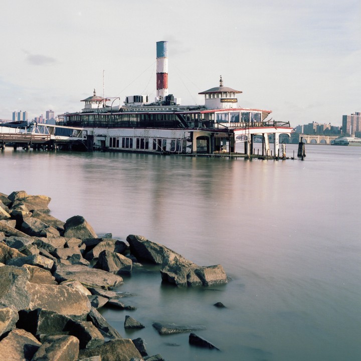 A large boat with a red, white, and blue smokestack is docked next to a rocky shore with a city skyline in the distance.