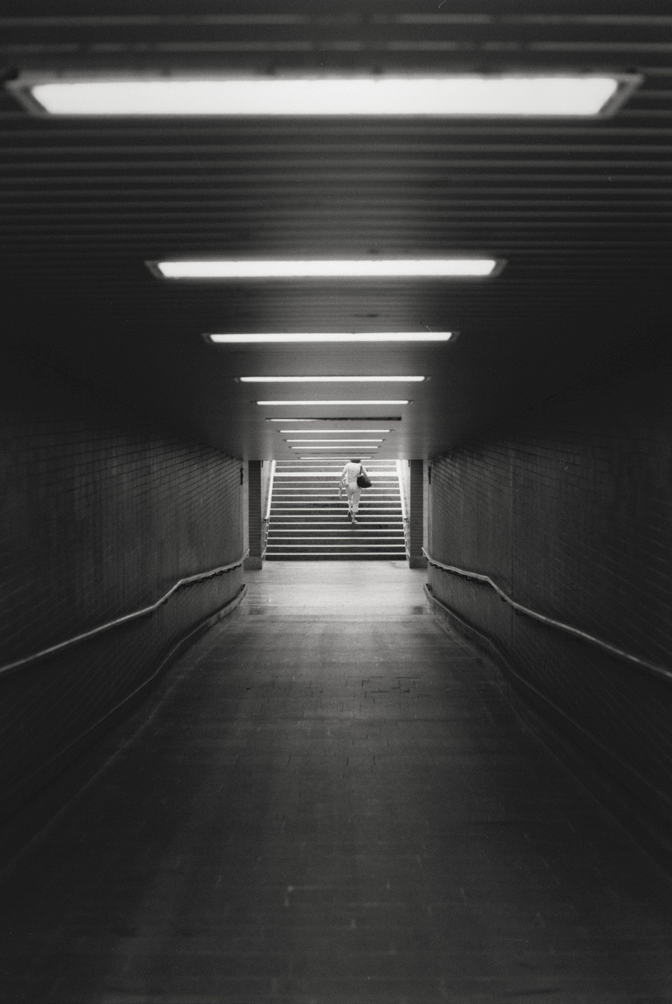 A person walks up a brightly lit staircase at the end of a dark, long underground tunnel.