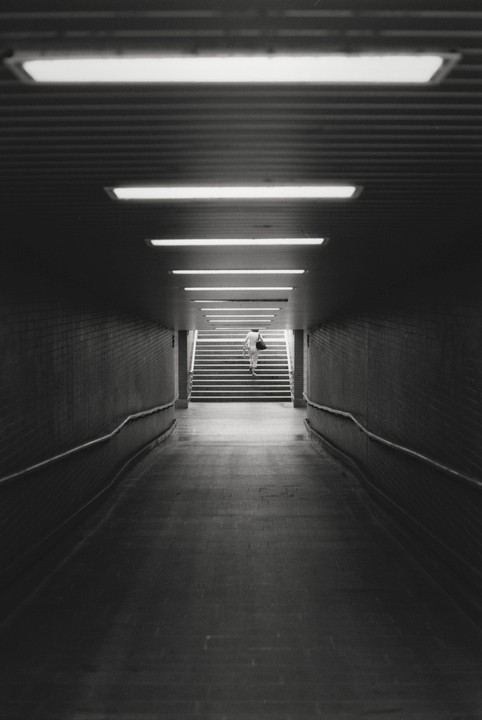 A person walks up a brightly lit staircase at the end of a dark, long underground tunnel.