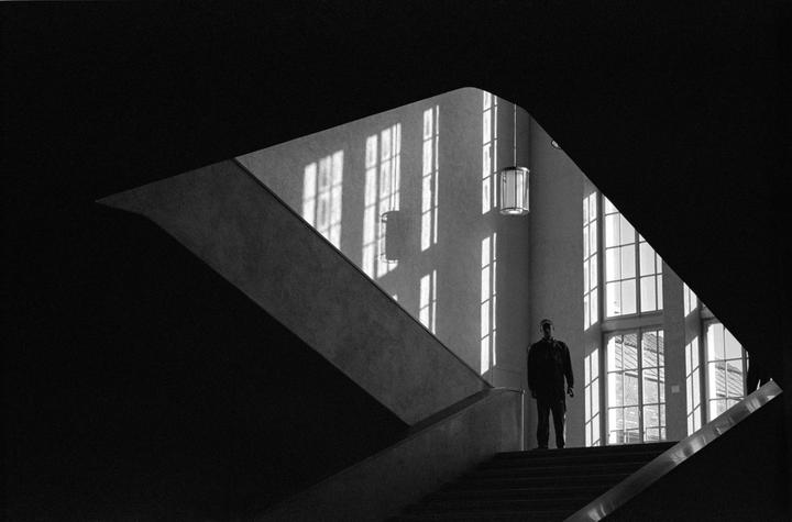 A man stands on a staircase with strong window light and shadows creating geometric patterns on the wall.