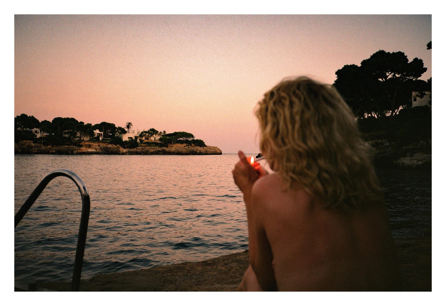 A woman with curly hair sits by the water at sunset, lighting a cigarette.