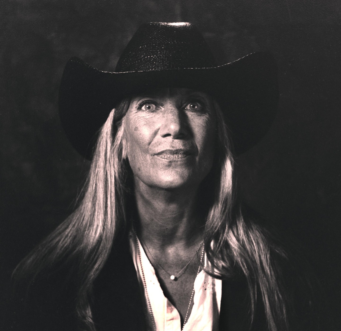 A woman wearing a cowboy hat and a necklace with a single pearl looks up at the viewer.
