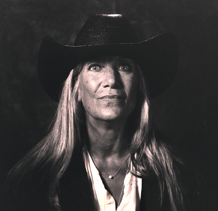 A woman wearing a cowboy hat and a necklace with a single pearl looks up at the viewer.