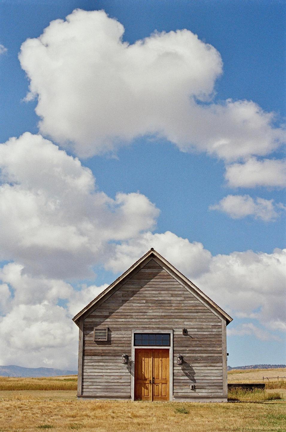 A wooden building with double doors stands in a field under a blue sky with white clouds.