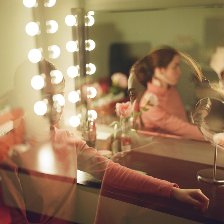 A woman sits in front of a vanity mirror with bright lights, creating a double exposure effect.