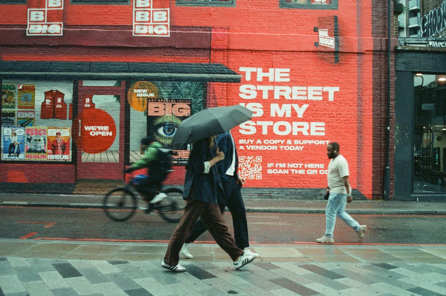 A person holding an umbrella walks past a red building with a mural that says "The Street is My Store" while a cyclist rides by.
