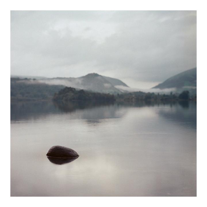A single rock sits in a still lake with mountains and fog in the background.