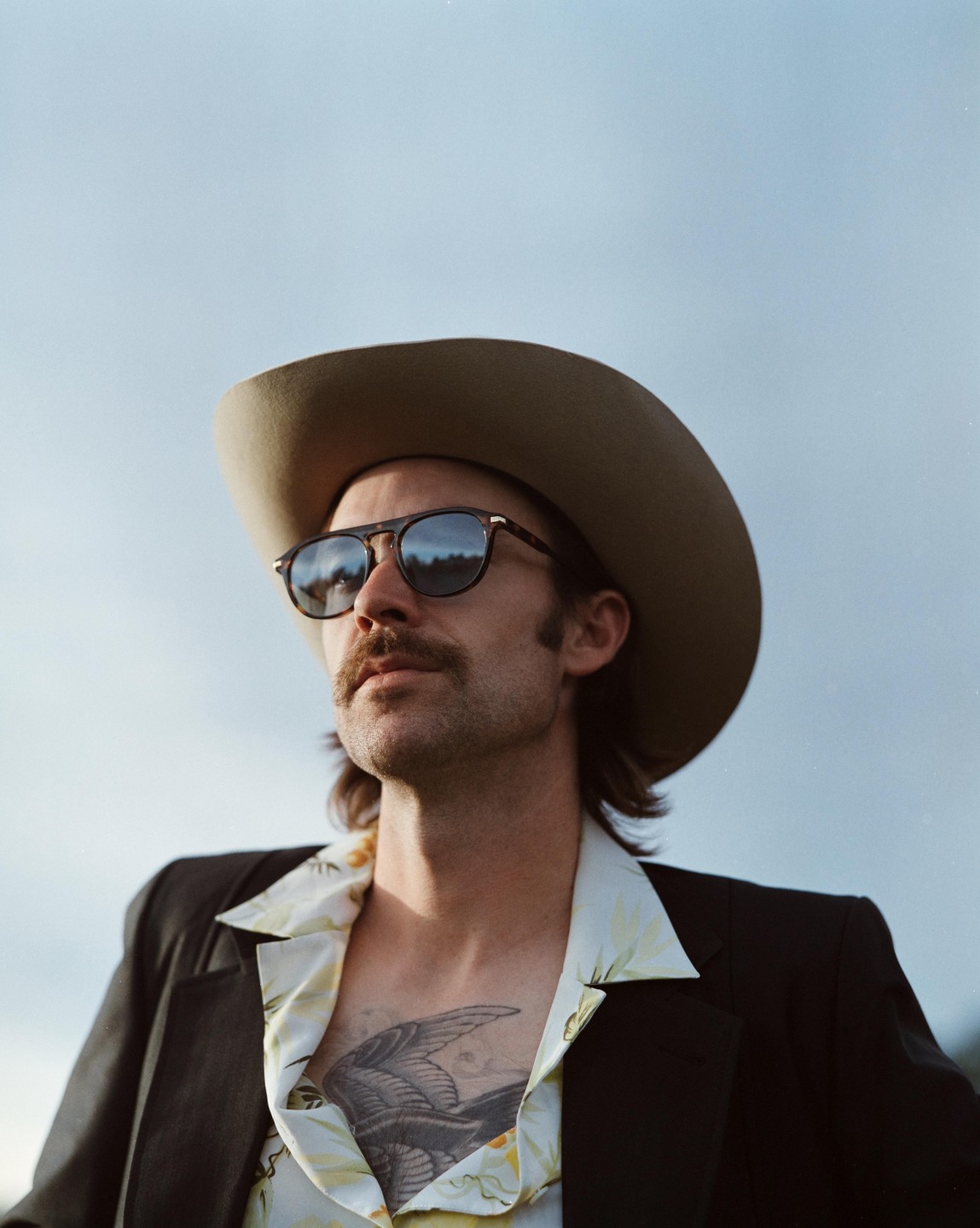 A man wearing a cowboy hat, sunglasses, and an open shirt revealing a chest tattoo looks up against a clear sky.