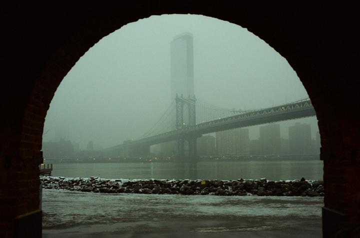 A view through an archway shows a bridge and a tall building across a river on a foggy day.