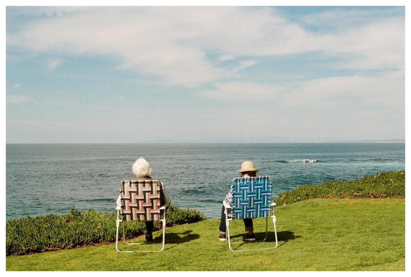 Two people sit in chairs on a grassy cliff overlooking the ocean.