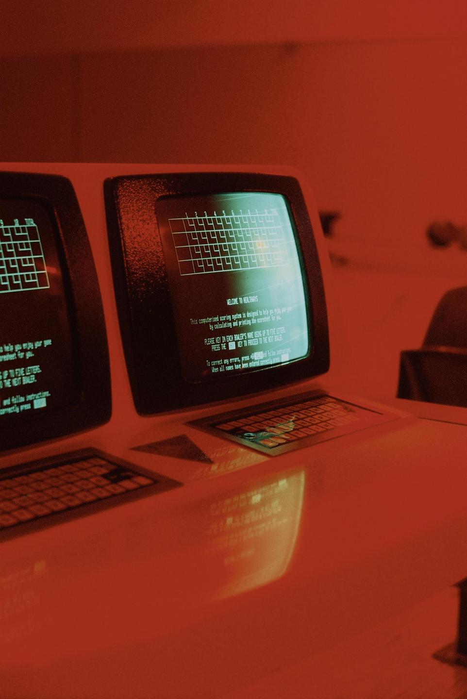 Two vintage computer monitors with keyboards are displayed on a reflective surface, showing text and a grid pattern on their screens.
