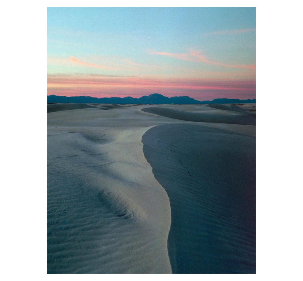 A vast desert landscape with sand dunes stretches towards mountains under a colorful sky.