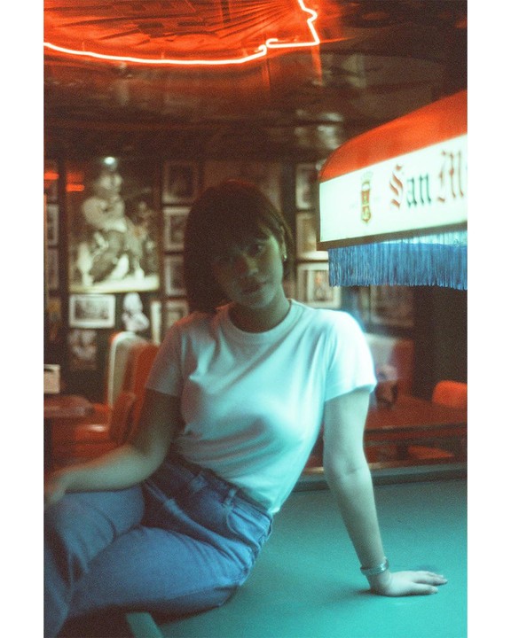 A woman in a white t-shirt and jeans sits on a pool table under a neon sign.