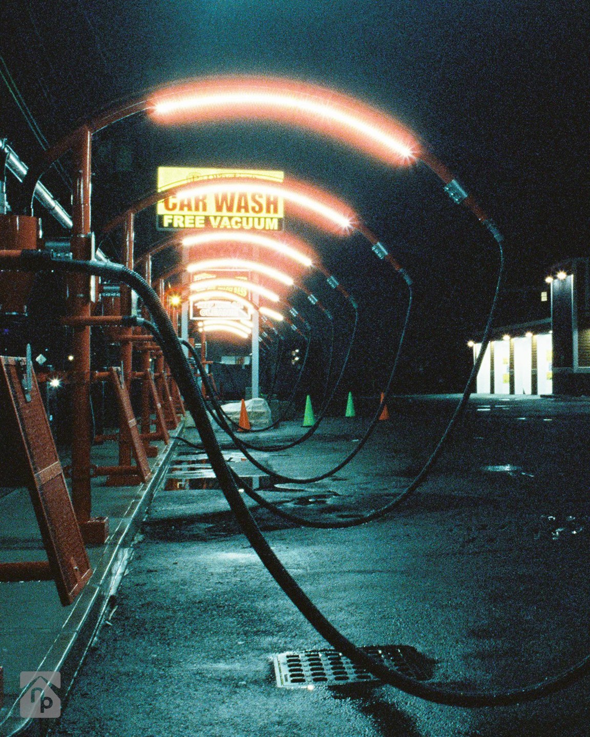 A car wash tunnel with glowing orange lights and hoses on the wet ground at night.