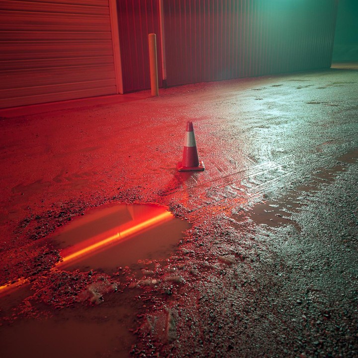 A traffic cone stands on a wet, gravelly surface with puddles reflecting red and green light.
