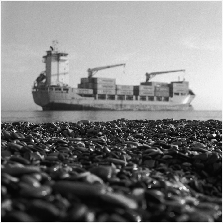 A cargo ship with containers is visible in the background, behind a foreground of pebbles on a beach.