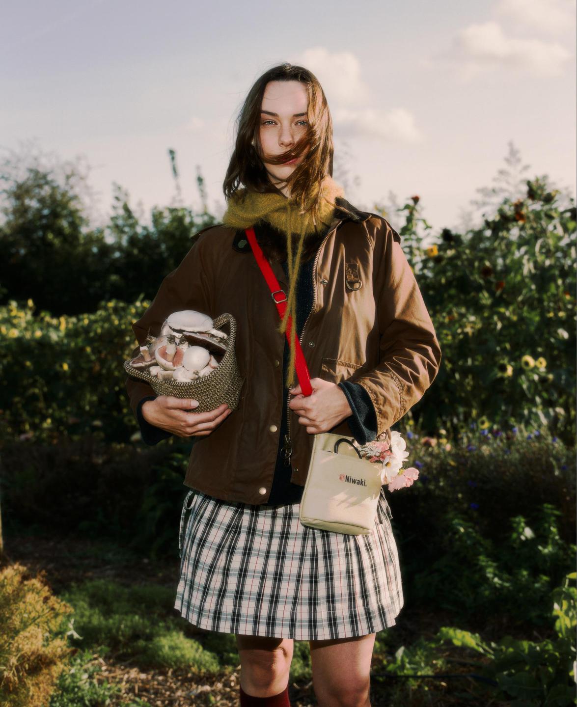 A woman stands in a garden holding a basket of mushrooms and a small bag with flowers.