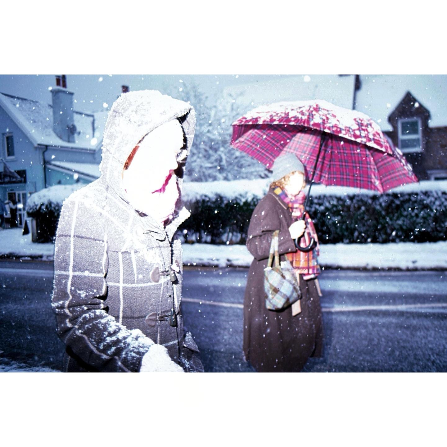 Two people walk on a snowy street, one holding a red umbrella.