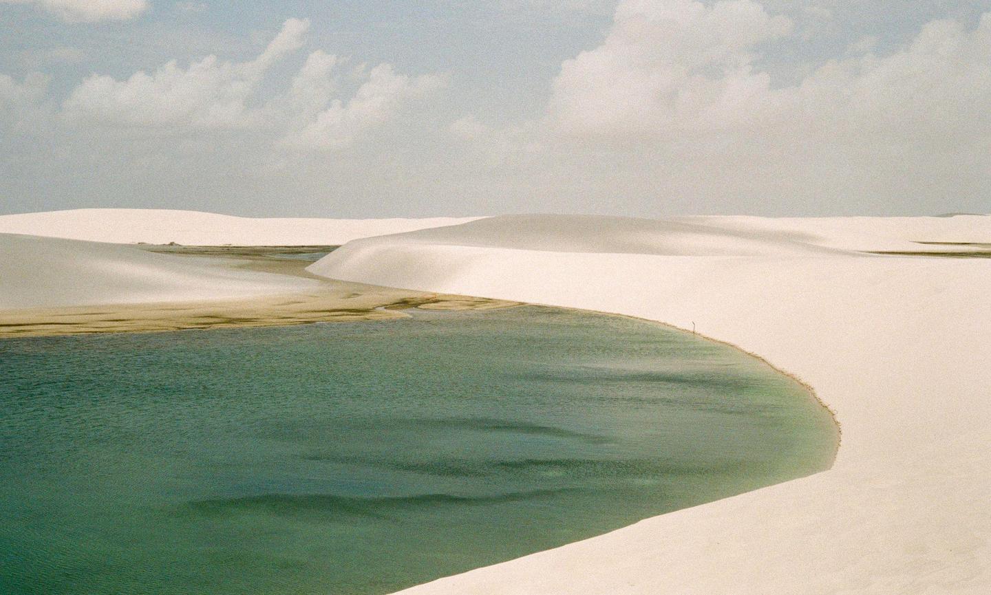 A body of water curves through a landscape of white sand dunes under a cloudy sky.
