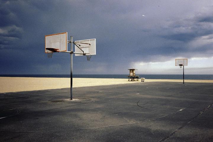 A basketball court sits empty on a sandy beach under a dark, cloudy sky with a lifeguard stand in the distance.