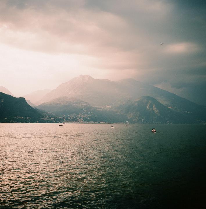 A body of water with several boats is surrounded by mountains under a cloudy sky.