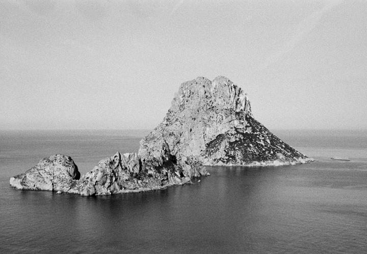 Two rocky islands rise from the sea under a bright sky, with a small boat visible in the distance.