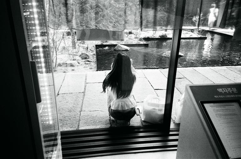 A woman with long hair crouches outside a window next to a body of water.