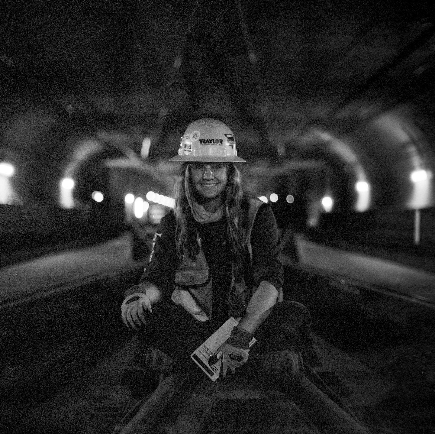 A woman wearing a hard hat and safety vest sits on a railroad track in a tunnel at night.
