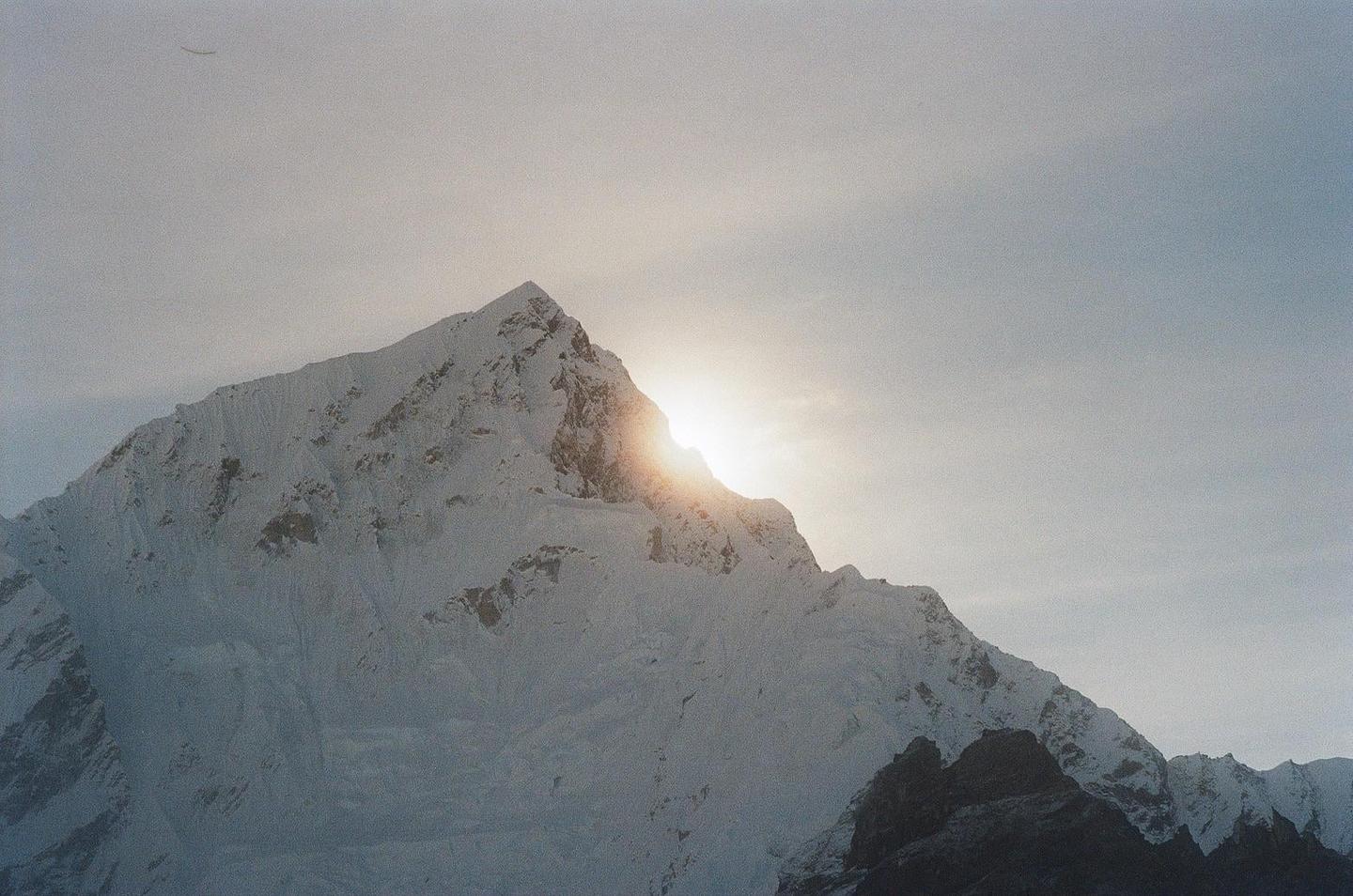 The sun shines brightly from behind a snow-covered mountain peak.
