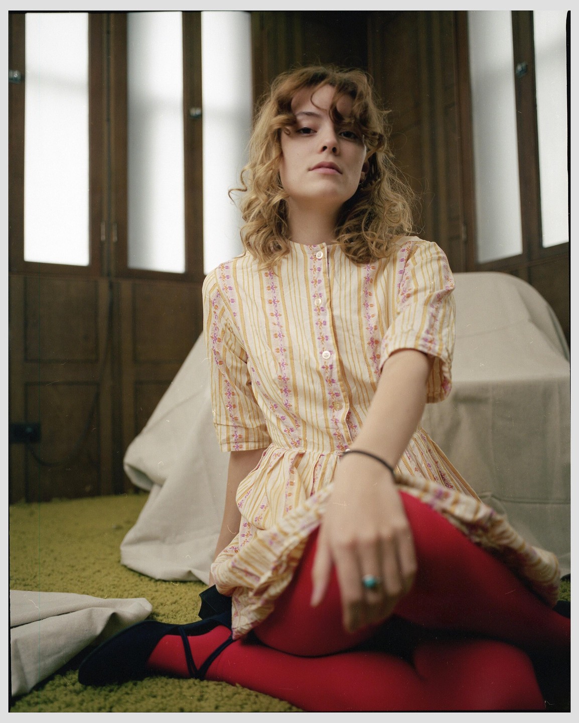 A woman with curly hair wearing a patterned dress and red tights sits on a green carpet.