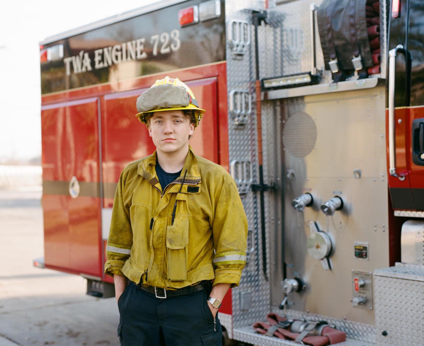 A man in a firefighter uniform and helmet stands in front of a fire truck.