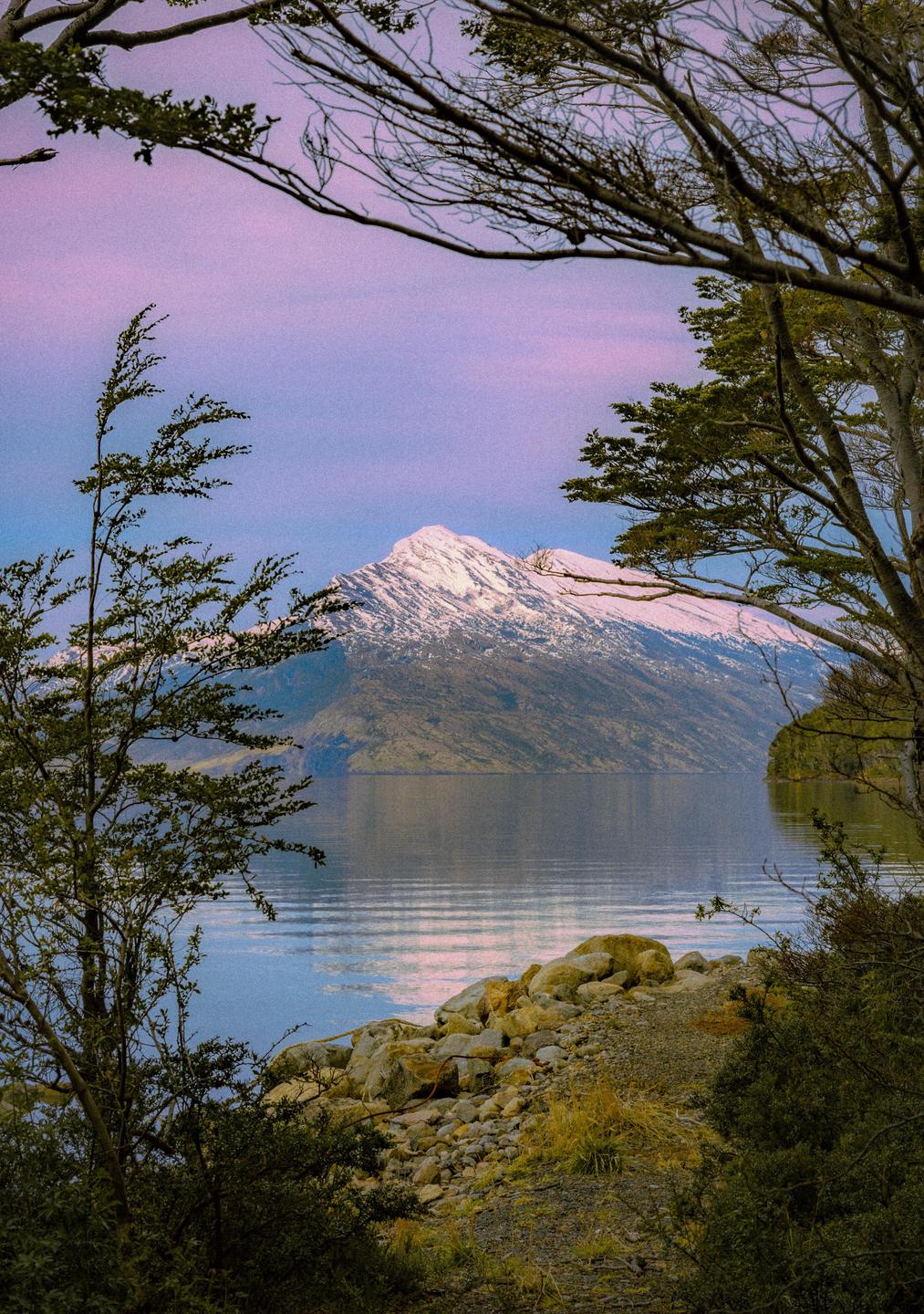 A snow-capped mountain rises above a lake, framed by trees and a rocky shore.