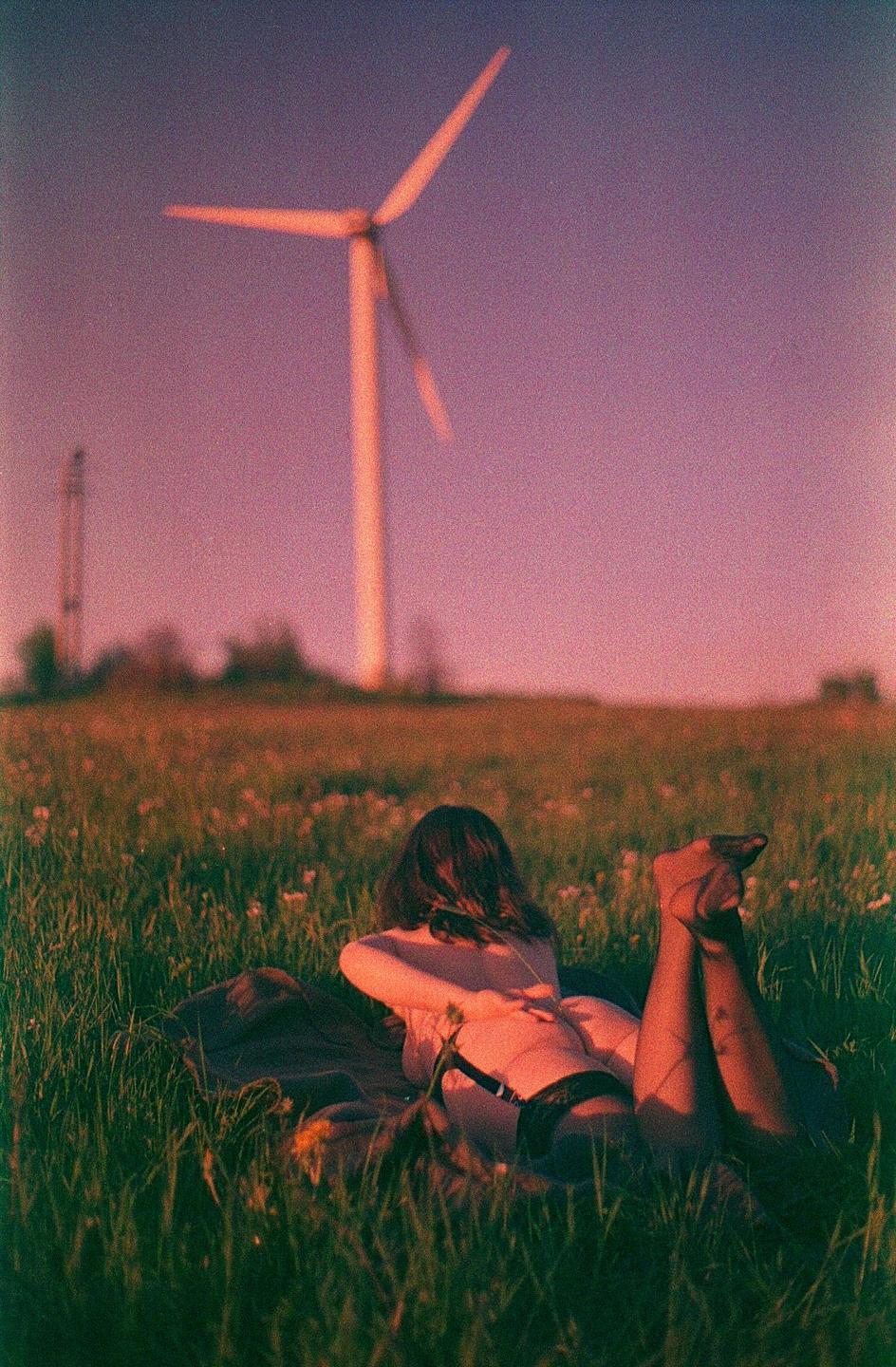 A woman lies in a field of grass with a wind turbine in the background at sunset.