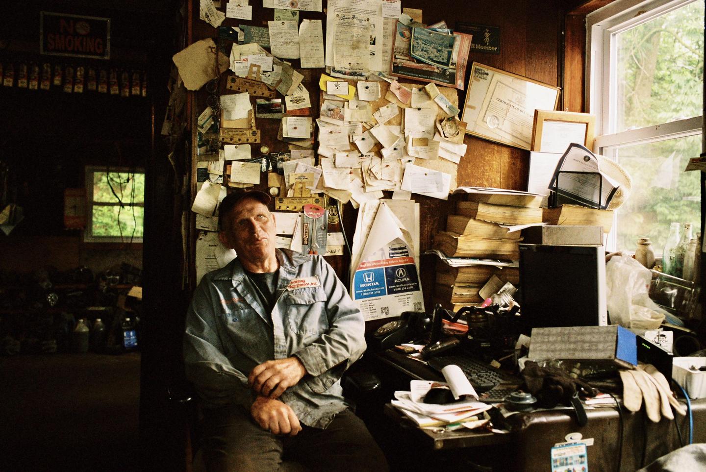 A man sits at a cluttered desk in a room with a wall covered in papers and two windows.