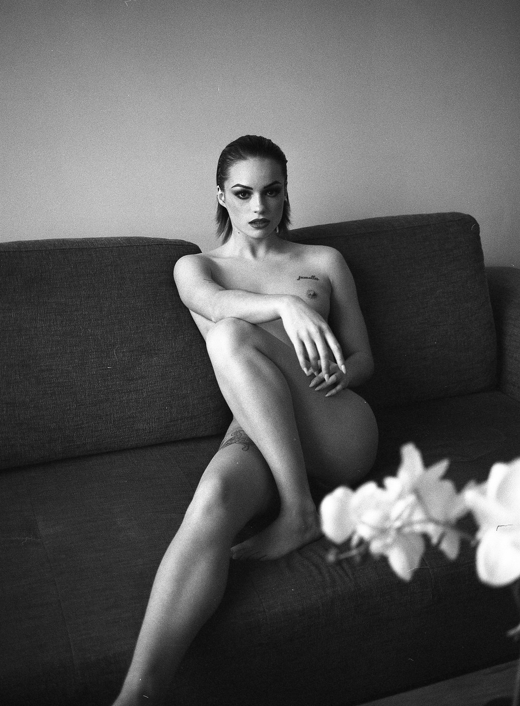 A nude woman with dark hair and makeup sits on a sofa with her legs crossed, looking directly at the viewer.