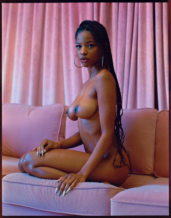 A topless woman with long braids and hoop earrings sits on a pink couch in front of pink curtains.