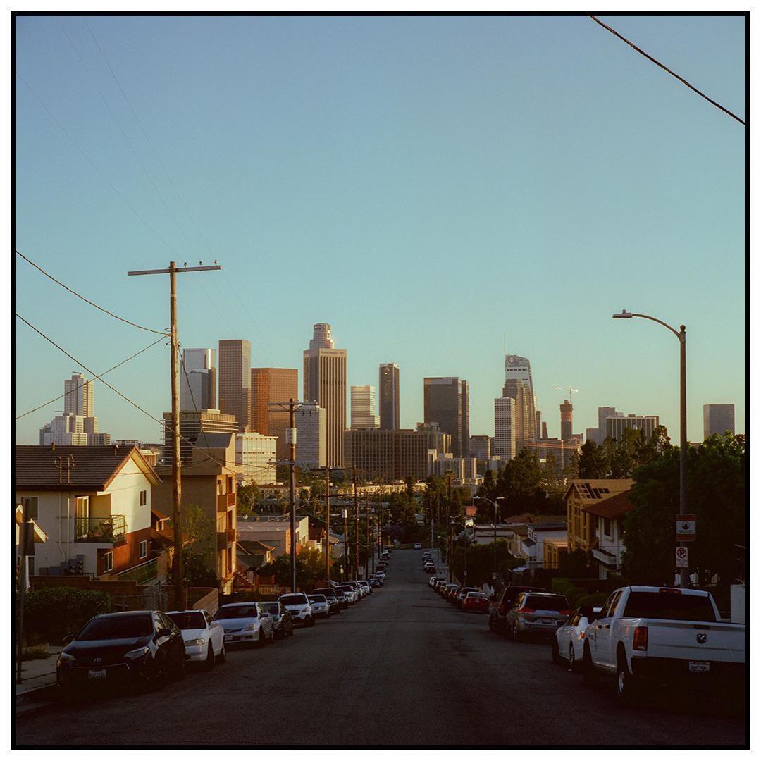 A street lined with parked cars leads to a distant city skyline under a clear sky.