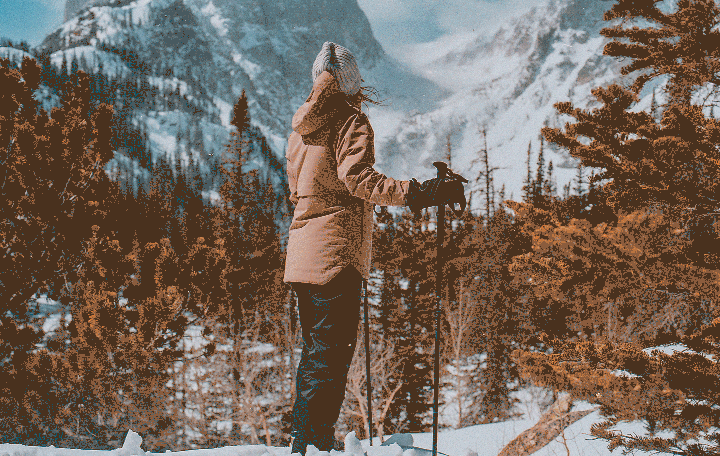 A person stands in a snowy mountain landscape, holding ski poles and looking out at the view.