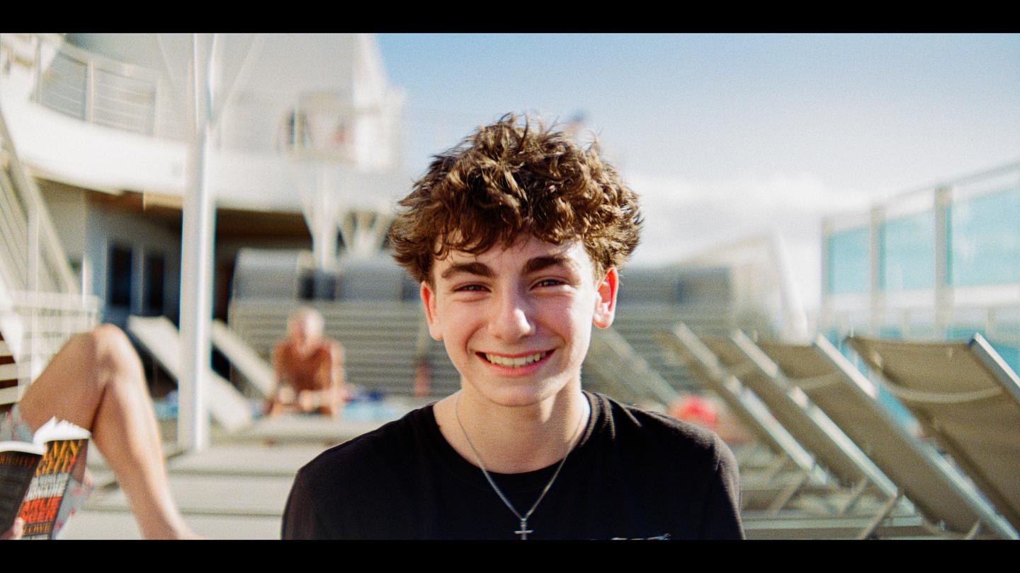 A young man with curly hair smiles at the camera while sitting outdoors on what appears to be a cruise ship deck.