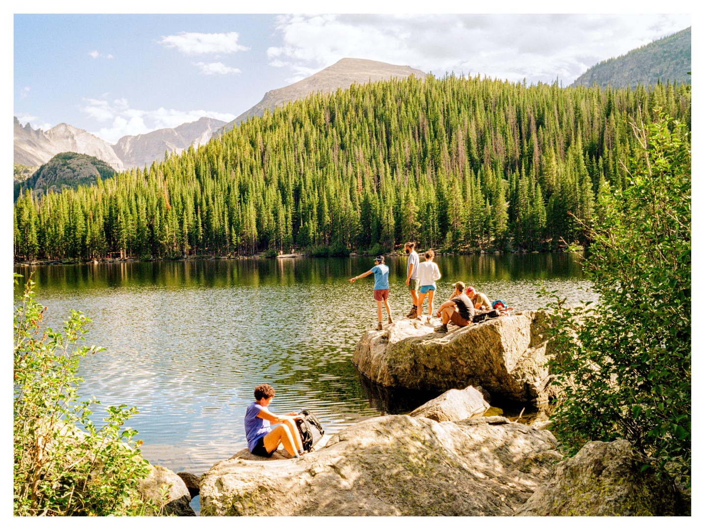 A group of people are gathered on rocks by a lake surrounded by a dense forest and mountains.