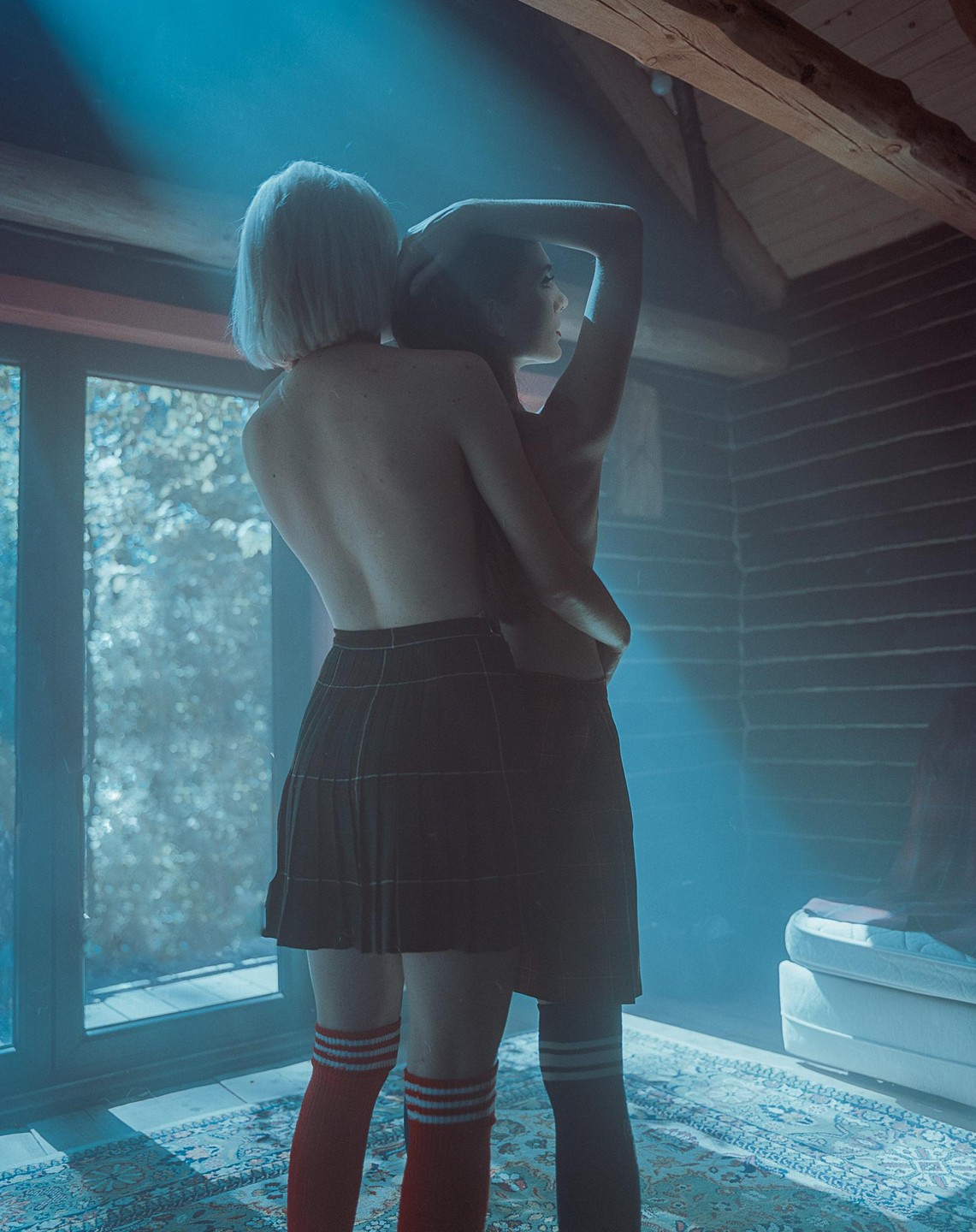 Two women embrace in a room with a window and a bed.