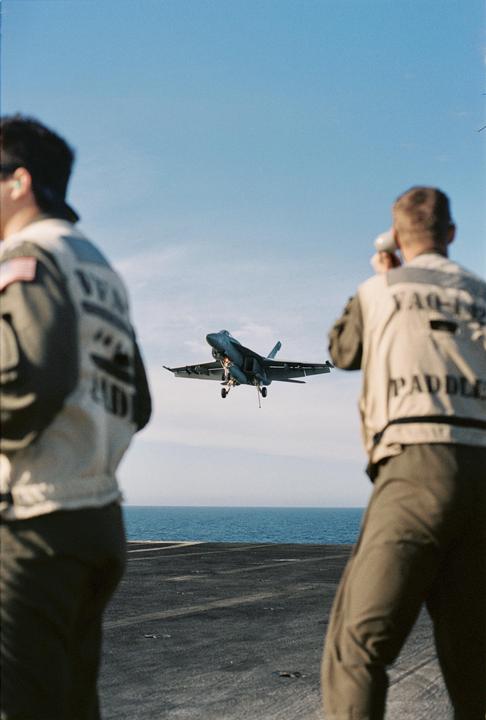 Two men stand on an aircraft carrier deck watching a jet aircraft approach for landing over the ocean.